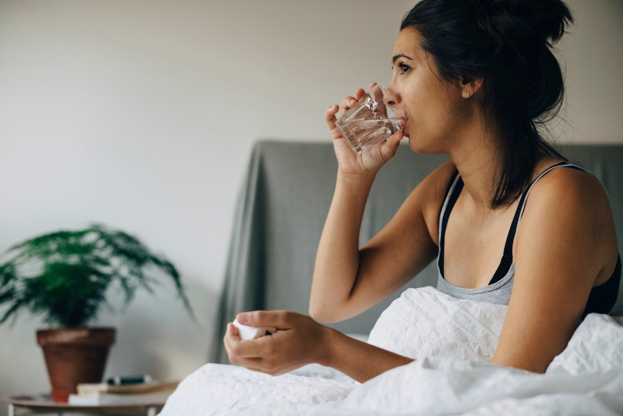 A woman drinking water to wash down a pill she's just taken while sitting in her bed