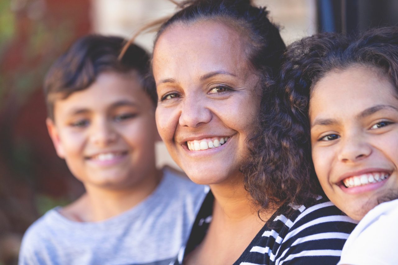 Aboriginal Family portrait with 1 parent and 2 children. They are sitting on the front porch. Everyone is happy and smiling. Could be a single mother.