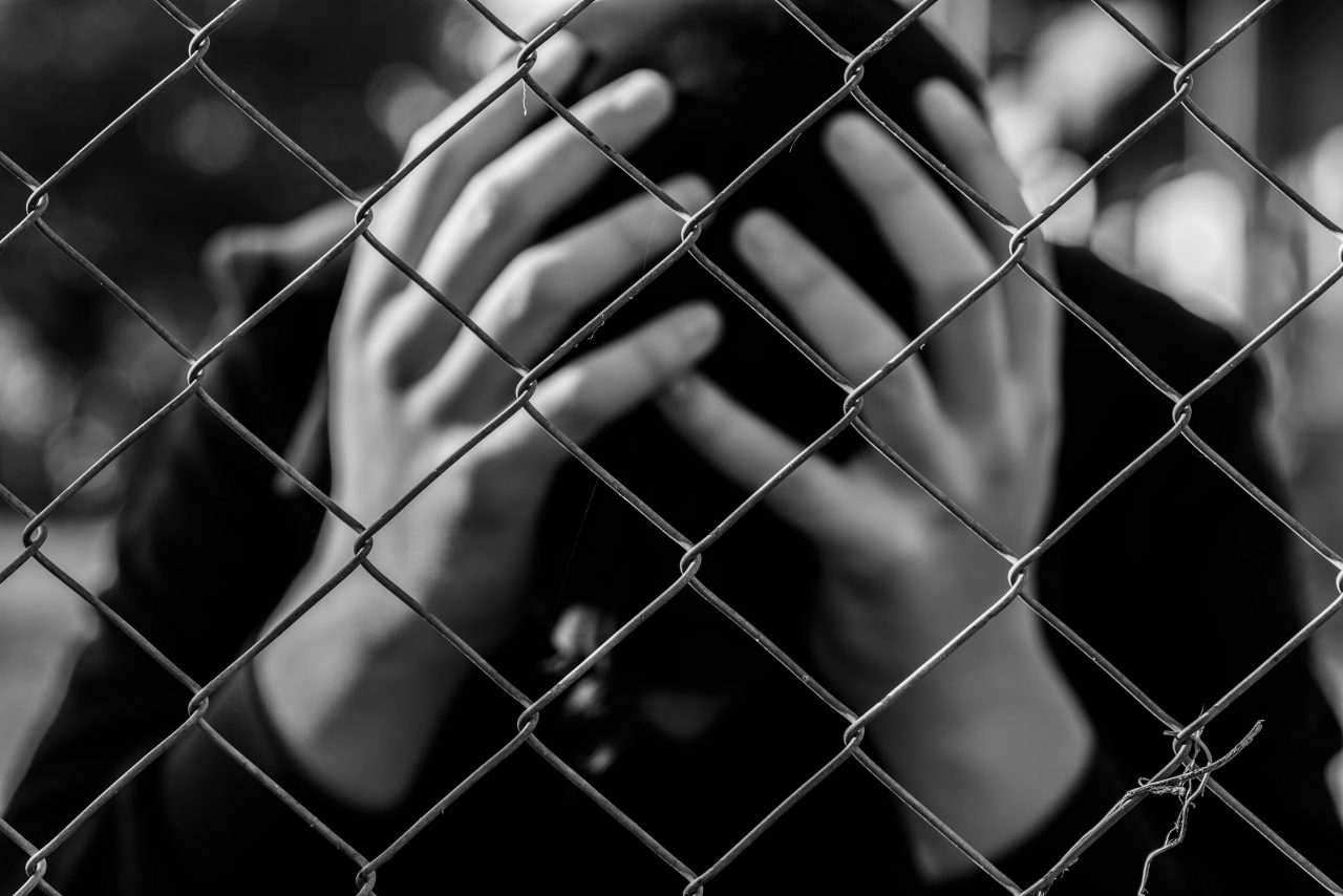 Young unidentifiable teenage boy holding hes head at the correctional institute in black and white, conceptual image of juvenile delinquency, focus on the wired fence.
