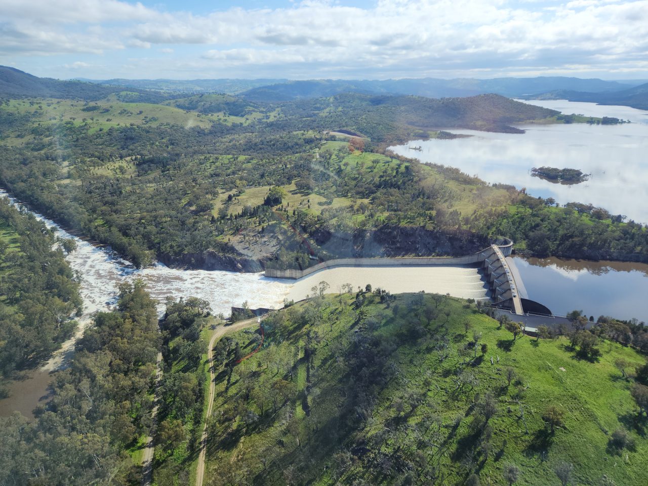 Aerial photo of a dam overflowing via the spillway into a river. The water is white where it is spilling out. The still water surface is reflecting the cloudy sky.  The land surrounding the dam is green and vegetated with trees but has some cleared land also very green. 