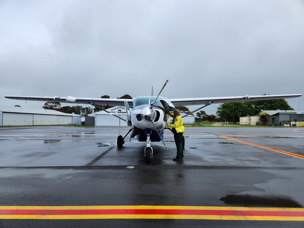 Photo of light aircraft on tarmac being checked by pilot. The sky is grey and the ground is wet