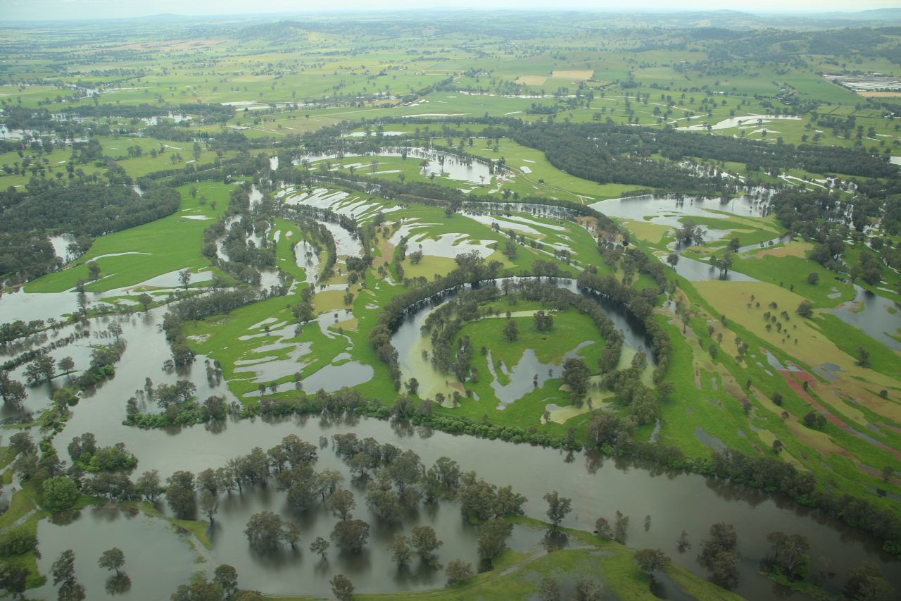 Aerial photo of river flooding beyond it's banks and across the floodplain.  Ground cover is very green with patches of trees around water courses have been inundated.