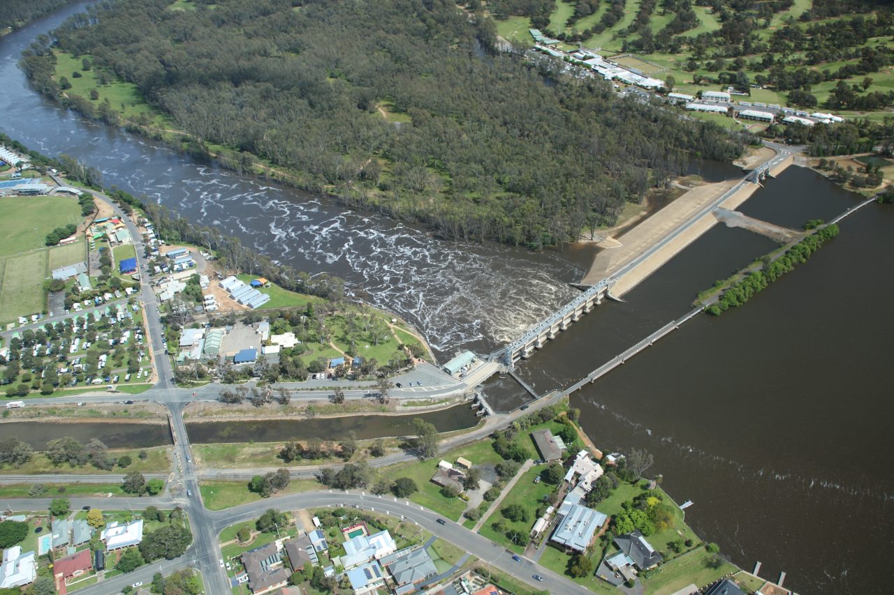 Aerial photo of lake and weir, water is flowing fast into the channel below the weir. .There is a town on one side of the channel and tree cover on the other side  followed by a resort and golf course. Bright sunny day.