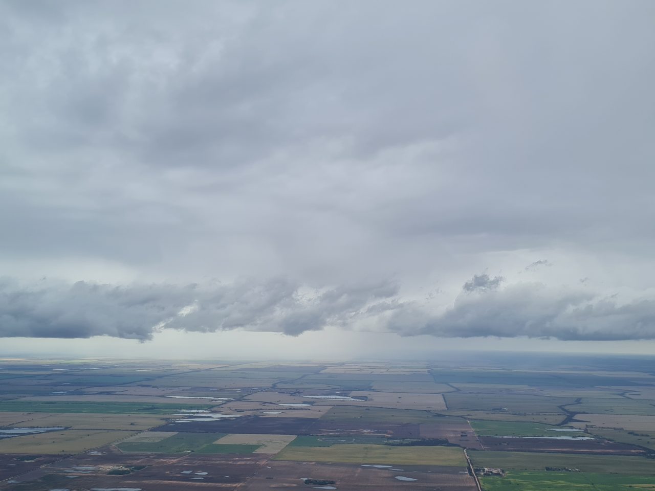 Aerial photo of grey cloud covered sky with a front of thick lower clouds in front of rain storms sitting on the horizon.  A patchwork of farm paddocks covers the  flat landscape with several smaller wetlands pooling in lower sections.