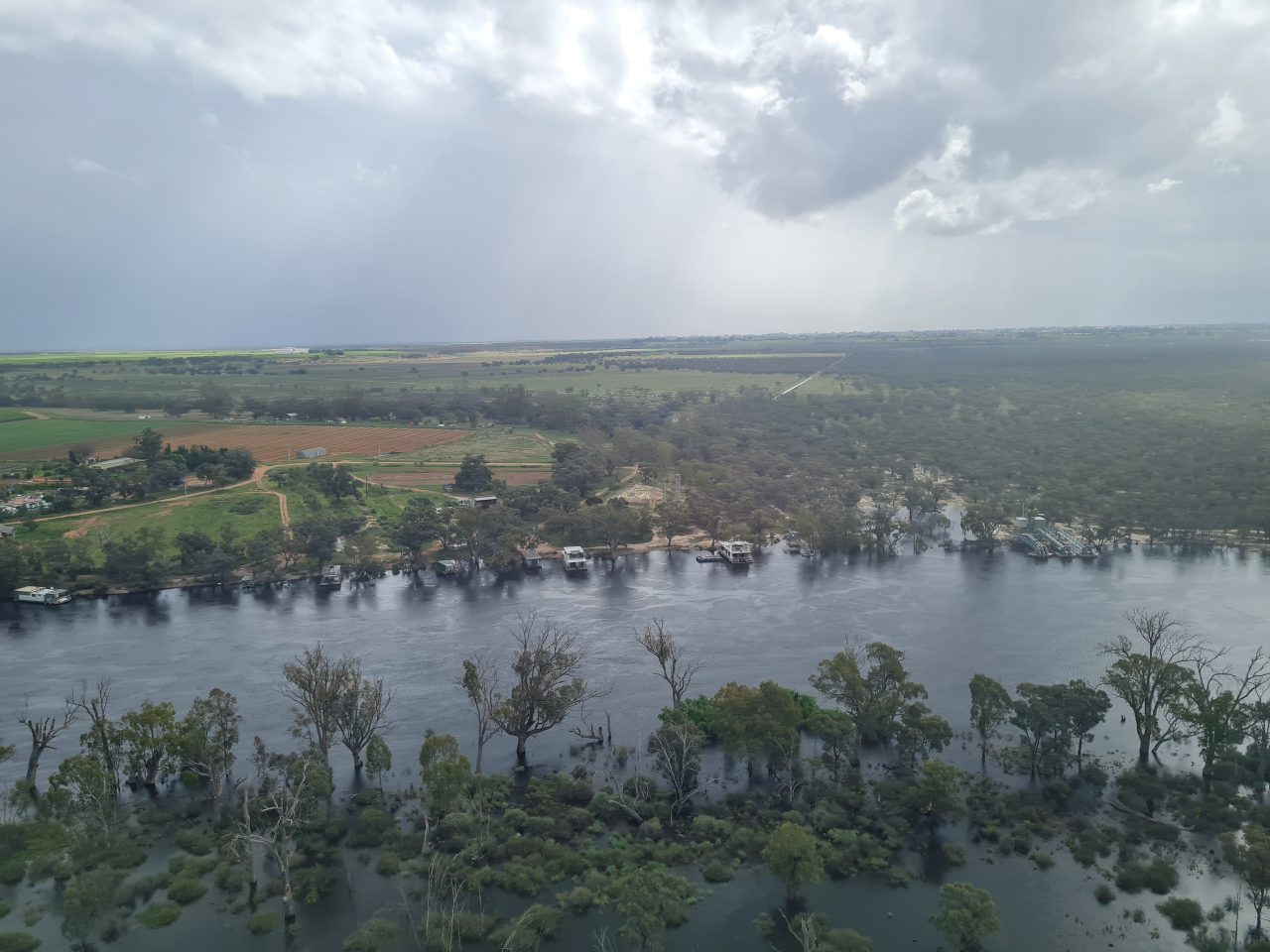 Aerial photo of a wide very full running river. The water is escaping the banks and inundating the surrounding floodplain. There are houseboats moored on the banks.  Farming paddocks and som patches of trees cover the floodplain. There is a dirt road with a house and other farm bulidings by the road. Sun is beginning to break through a cloudy sky.