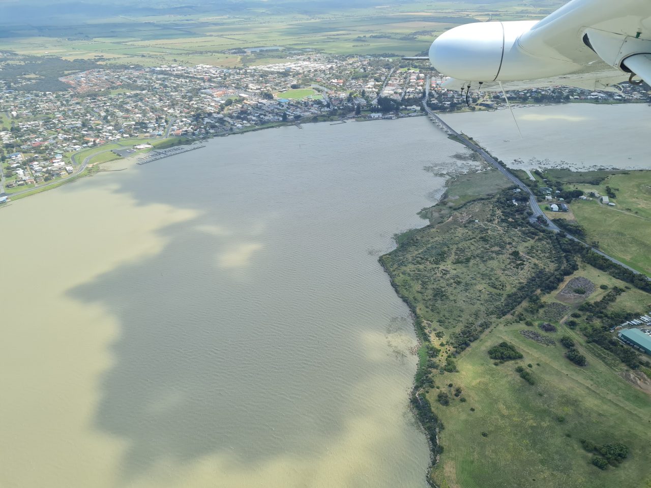 Aerial photo of a bend in a wide river, filled with light brown water.  The sky is not visible but shadows from the clouds are visible on the surface of the water. There is a bridge crossing the river to a town. Green farm paddocks surround the town. THe wing of the plane is in the top right corner of the photo.