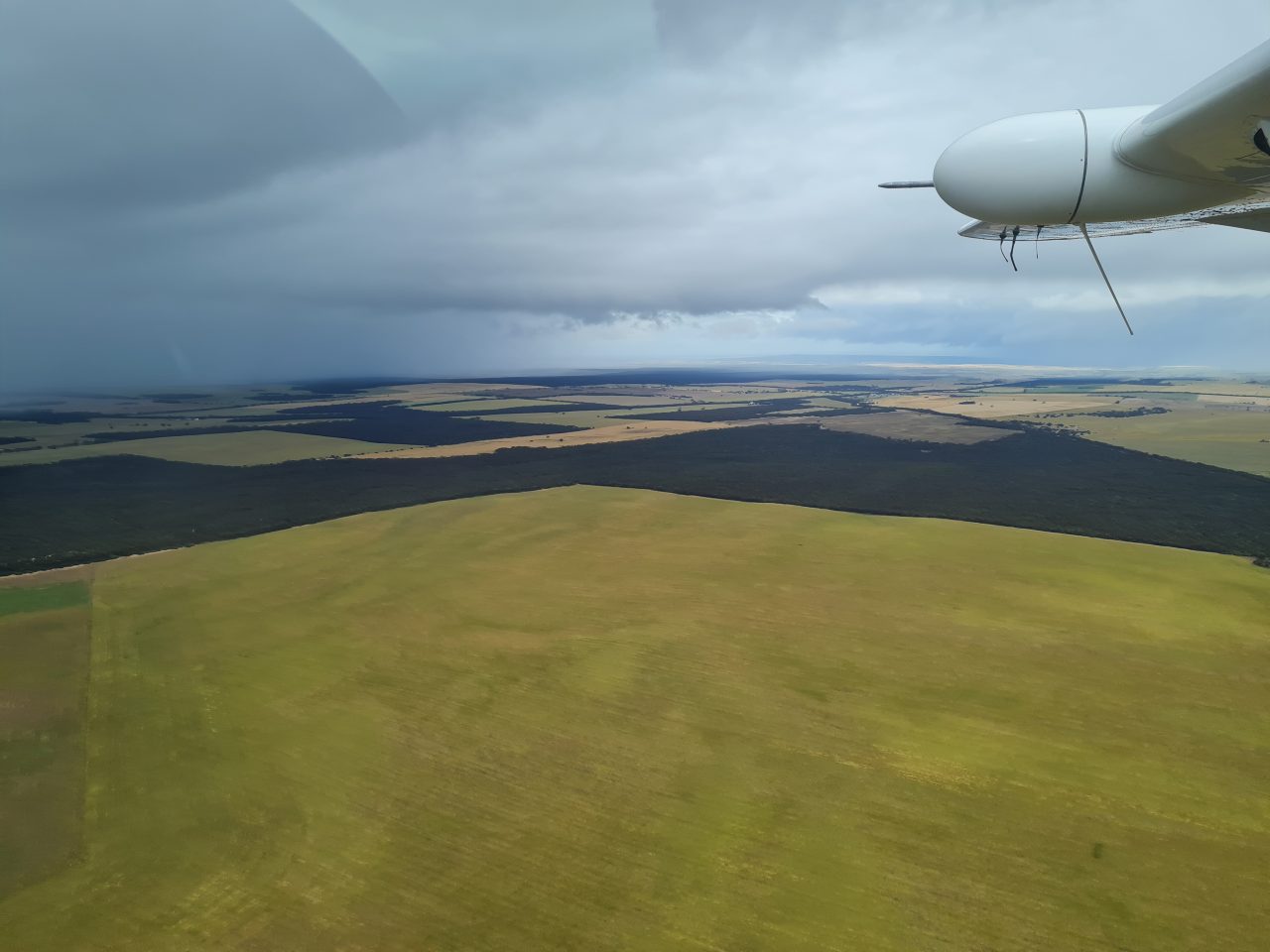 Aerial photo of clouds and rain of a storm front making their way across the land which is a patchwork of cleared and forested  areas. The plane wing is visible in the side of the picture.