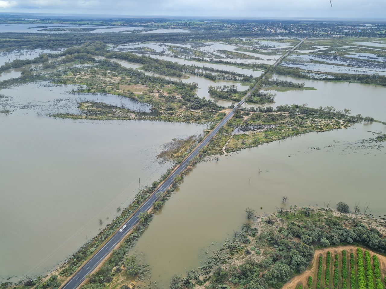 Aerial photo of floodwaters surrounding a long straight stretch of road that is just above the water level and leading toward a town in the distance.