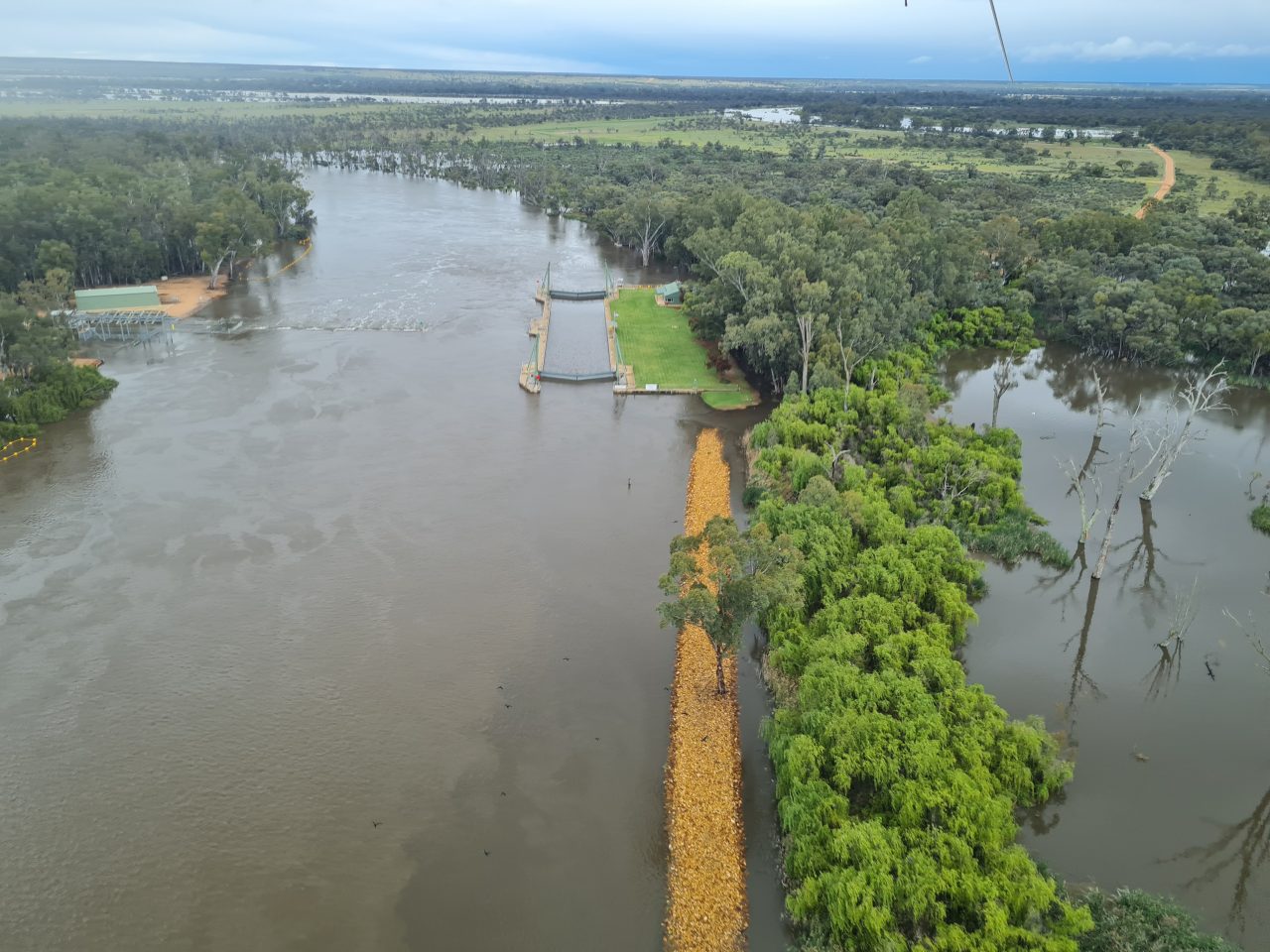 Aerial photo of a lock structure within the waters of a flooded river. THe clearing beside the lock is only just above the water level. The river is spreading beyond the tree lined banks and across the floodplain. 
