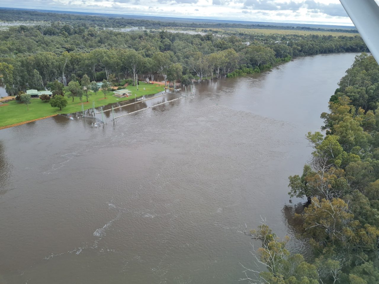 Aerial photo of a lock structure within a high running river. The river is flooding beyond the tree lined banks.  There is a clearing with several buildings that is only just above the water level.
