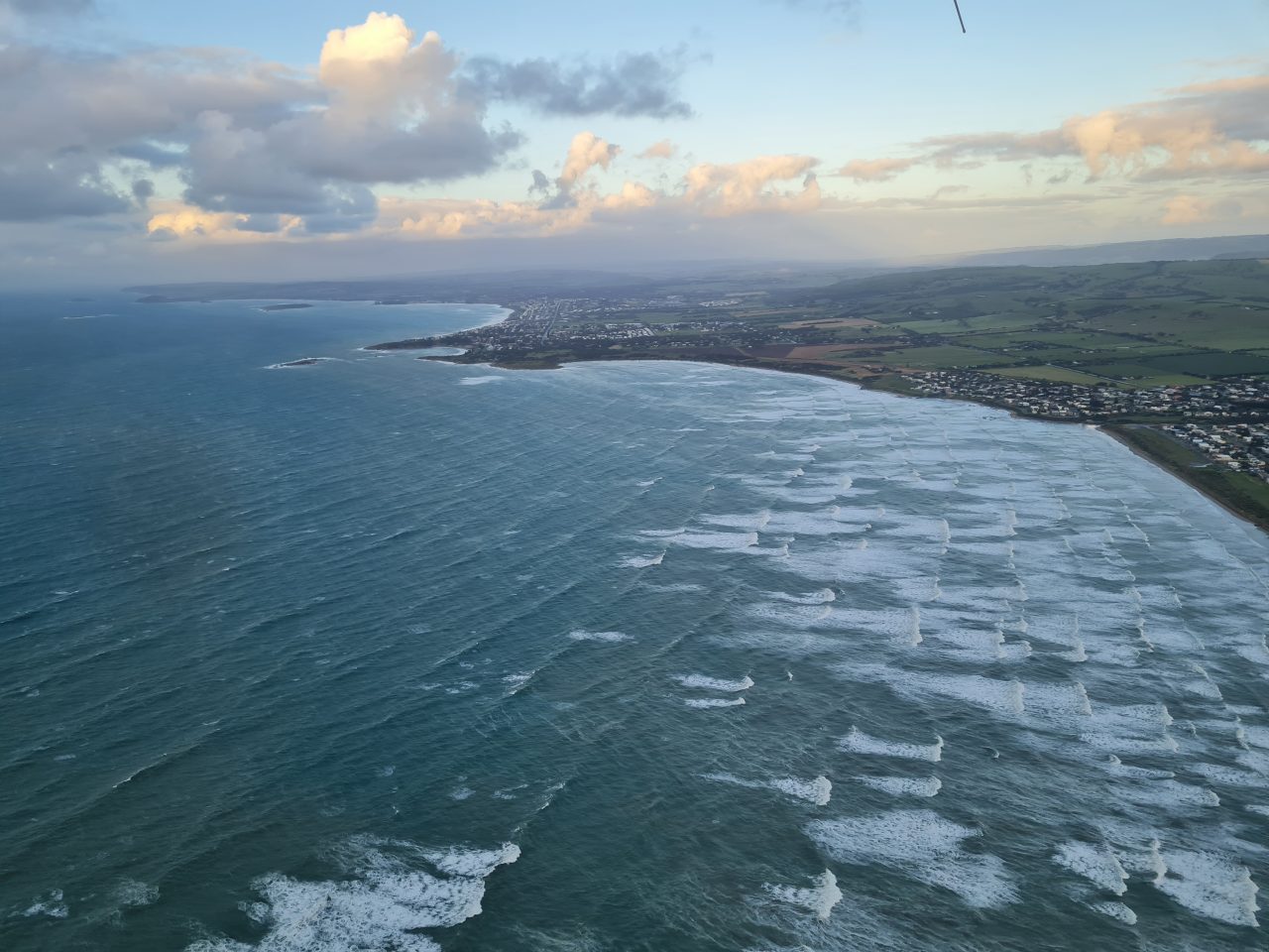 Aerial photo of coastline, ocean and land. Waves are breaking far out into the bay indicating a shallow coast. There are towns on the points on the coast. The sun is shining on the clouds  and there are patches of clear sky.