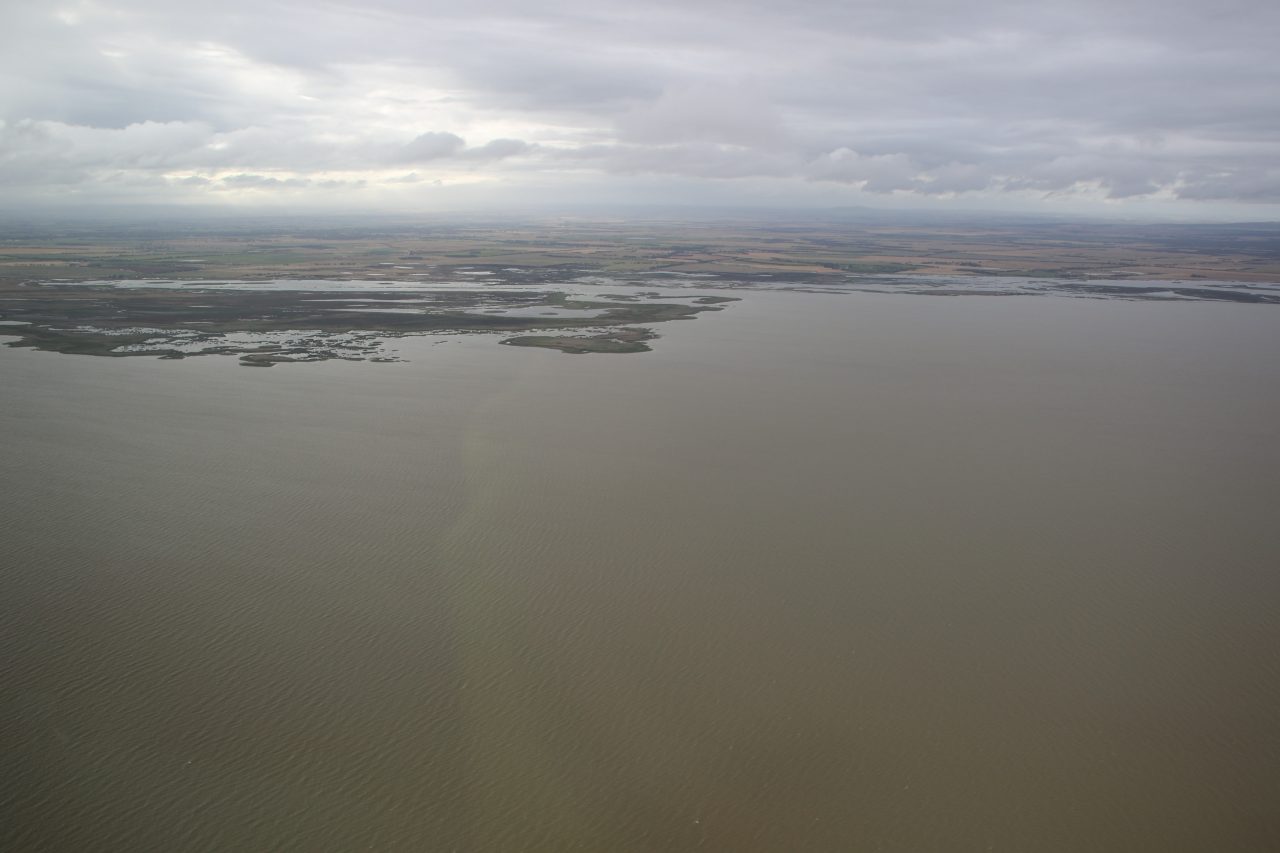 Aerial photo of part of a large lake filled with brown water and the surrounding landscape under a grey cloudy sky