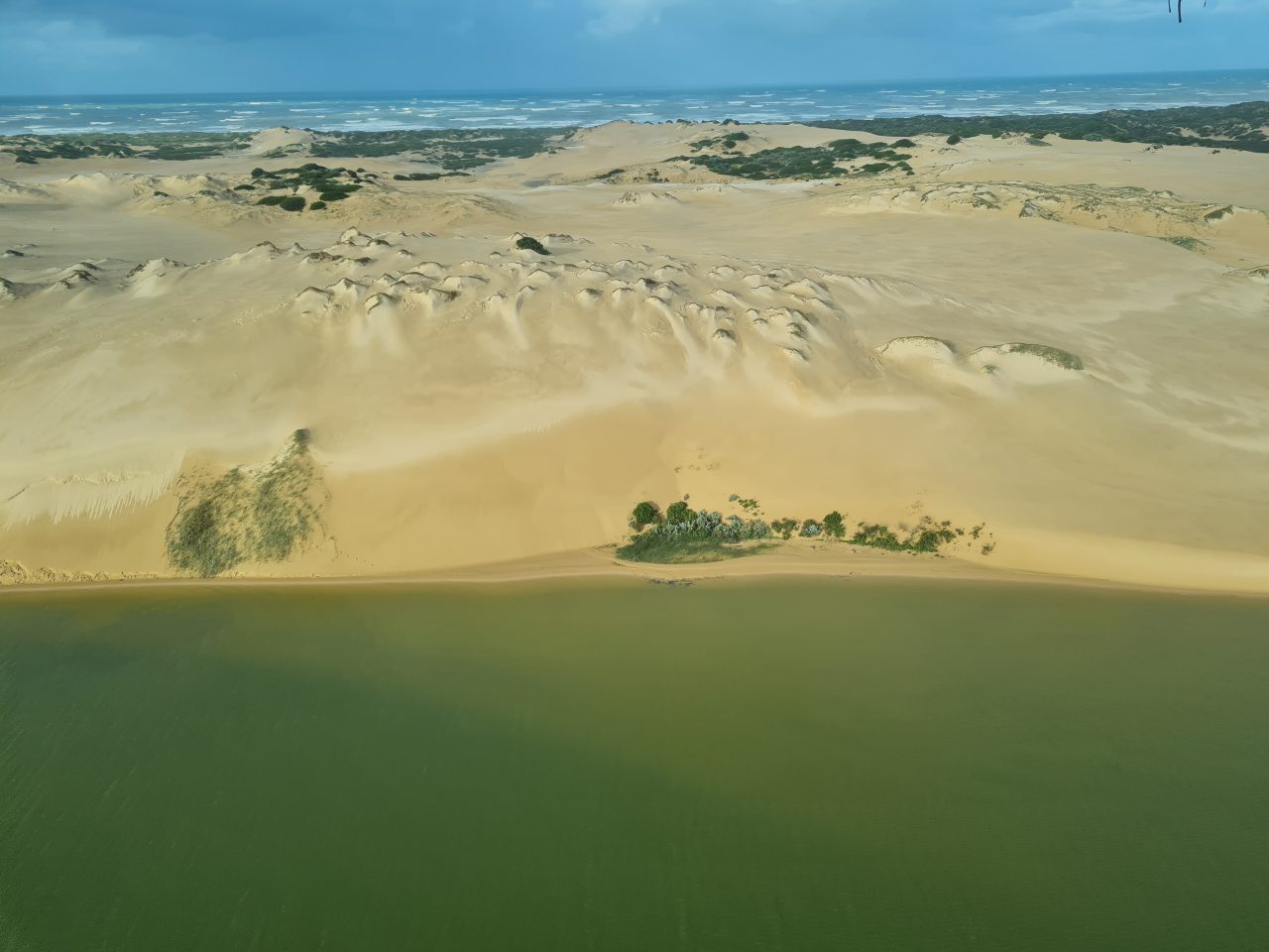 Aerial photo of coastal lake edge consisting of a series of sand dunes with the choppy ocean in the background. 