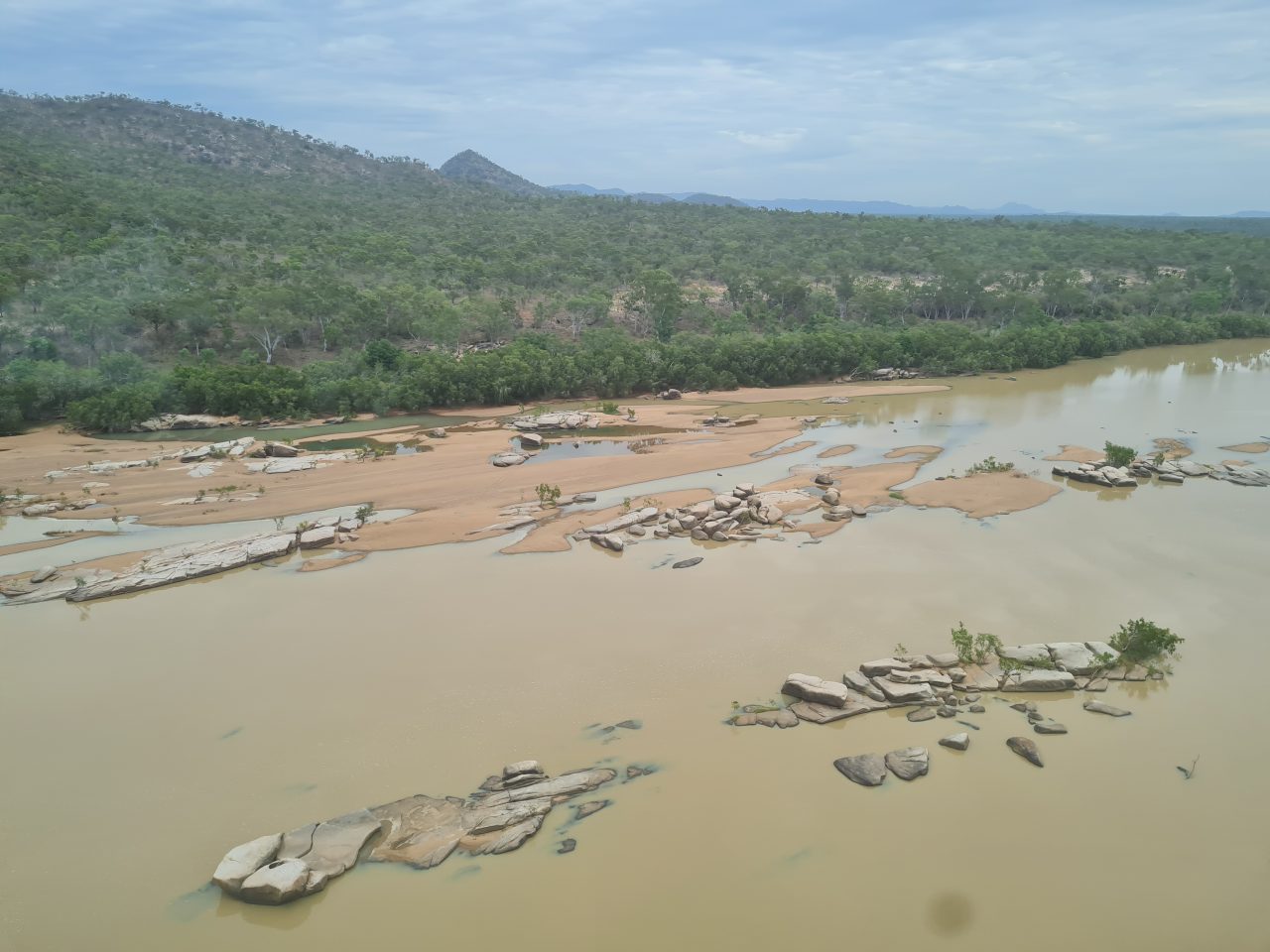 Aerial photo of river with brown water, sand formations and rocky outcrops running next to a naturally vegetated hill. Mountains in the background and on a cloudy day.