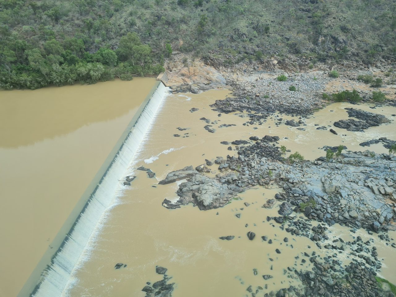 Aerial photo of  weir running across river, flowing with yellow-brown water and exposed rocks on the lower side of weir
