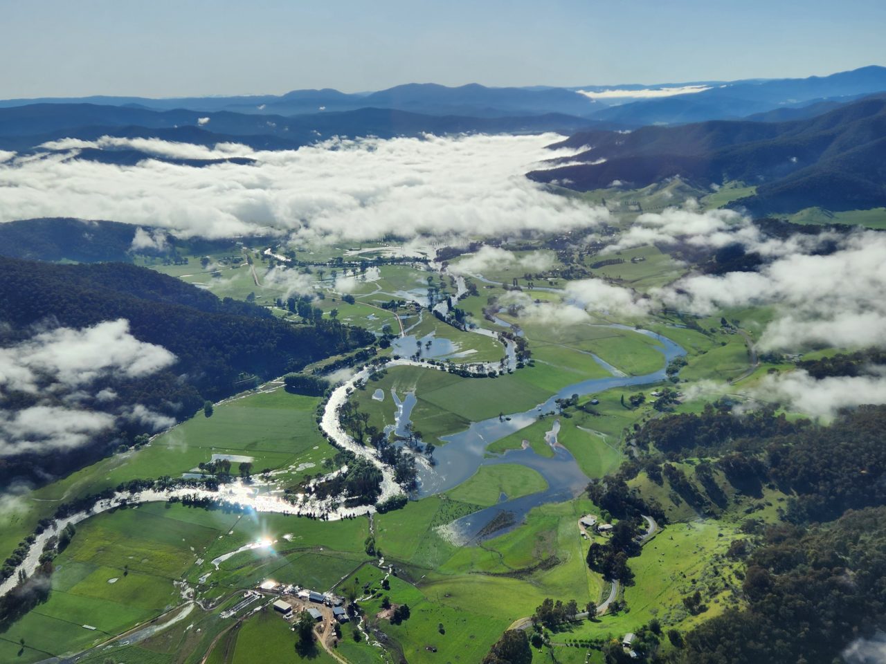 High aerial photo of a river meandering across a green cleared floodplain. There are densely forested hills and mountains extending up to the horizon. 