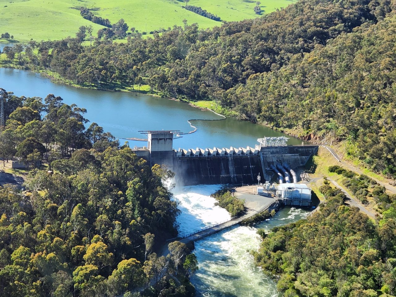 Aerial photo of a dam wall that has water spilling over. Surrounding hills are covered in trees with green paddocks in the background .