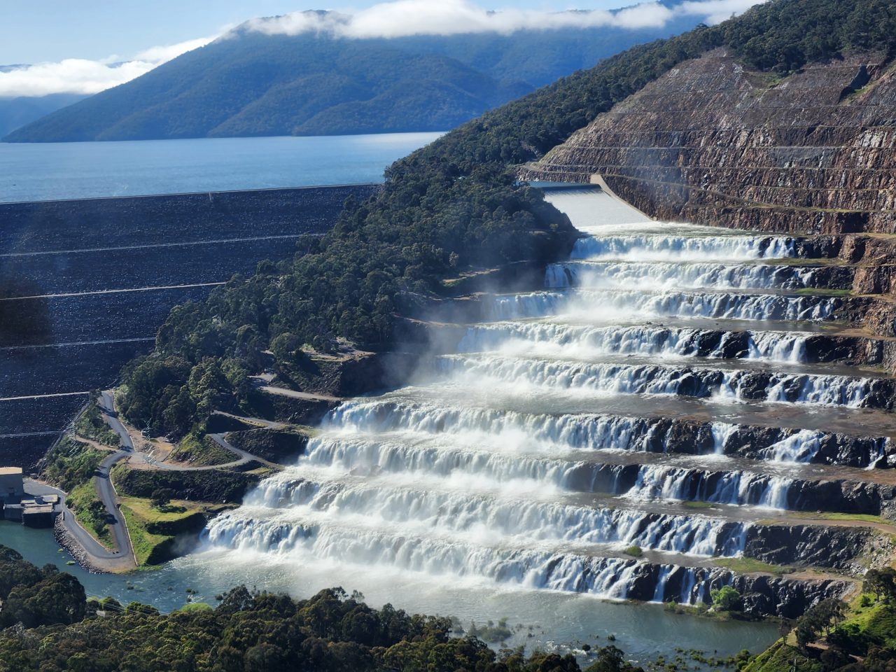 Low aerial photo of a terraced dam spillway covered in white water gushing down like a waterfall into the river below.