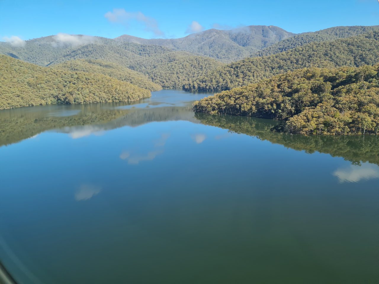 Low level aerial photo of a very full dam surrounded by densely tree covered hills rising into mountains on a sunny day with a  couple of low clouds causing the water to reflect the sky. 