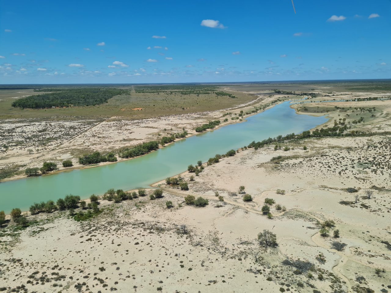 Aerial photo of a lake with light aqua water surrounded by light yellow sand  sporting sparse vegetation that is  more concentrated around the edge of the lake. The landscape is flat.