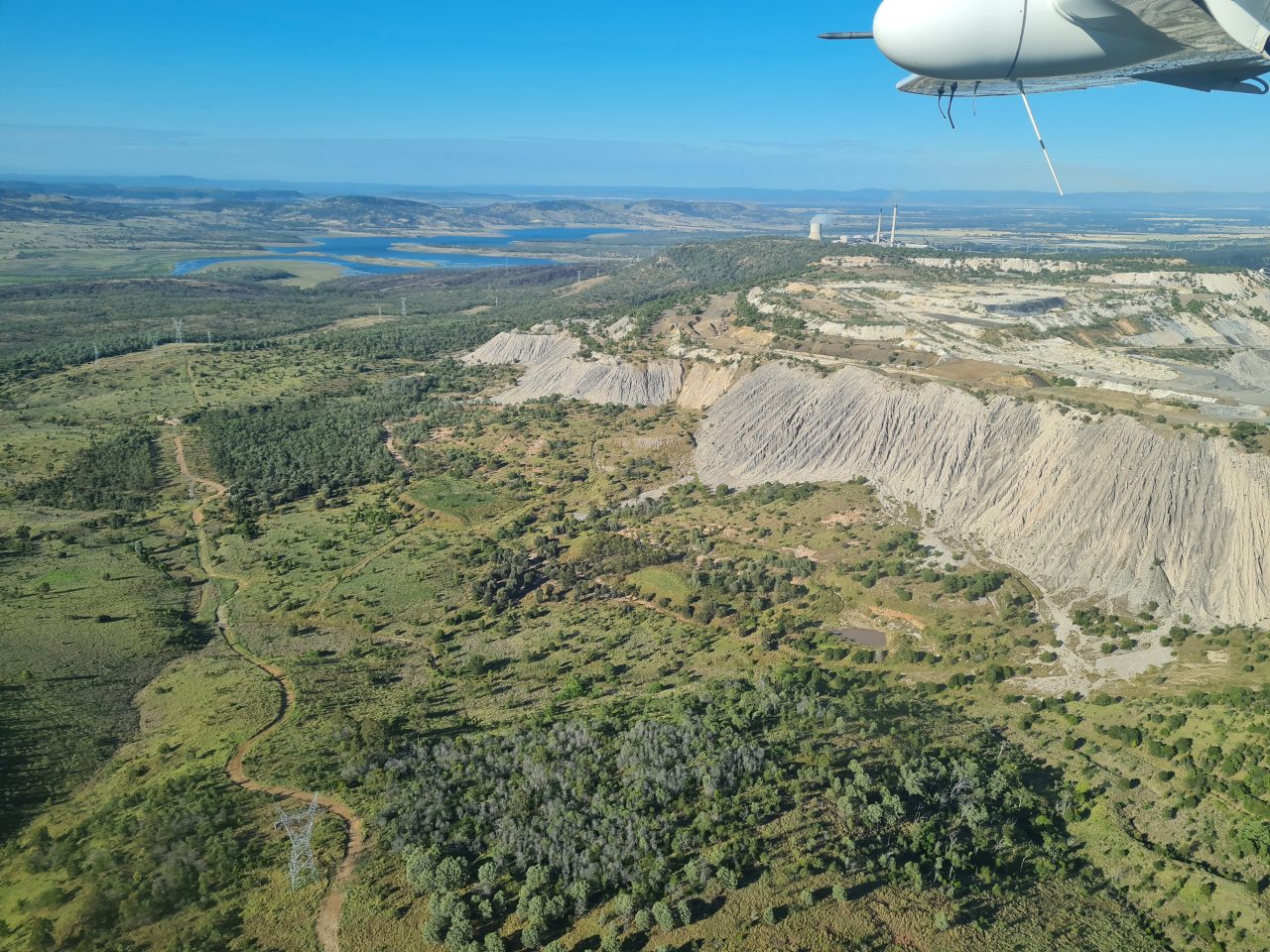 Aerial photo of open cut coal mine and coal fired power station and it's large blue cooling dam in the background. In the foreground is a green vegetated rolling landscape with several dirt roads cut out on ridges leading to the power station lined by power transmission towers and powerlines. 