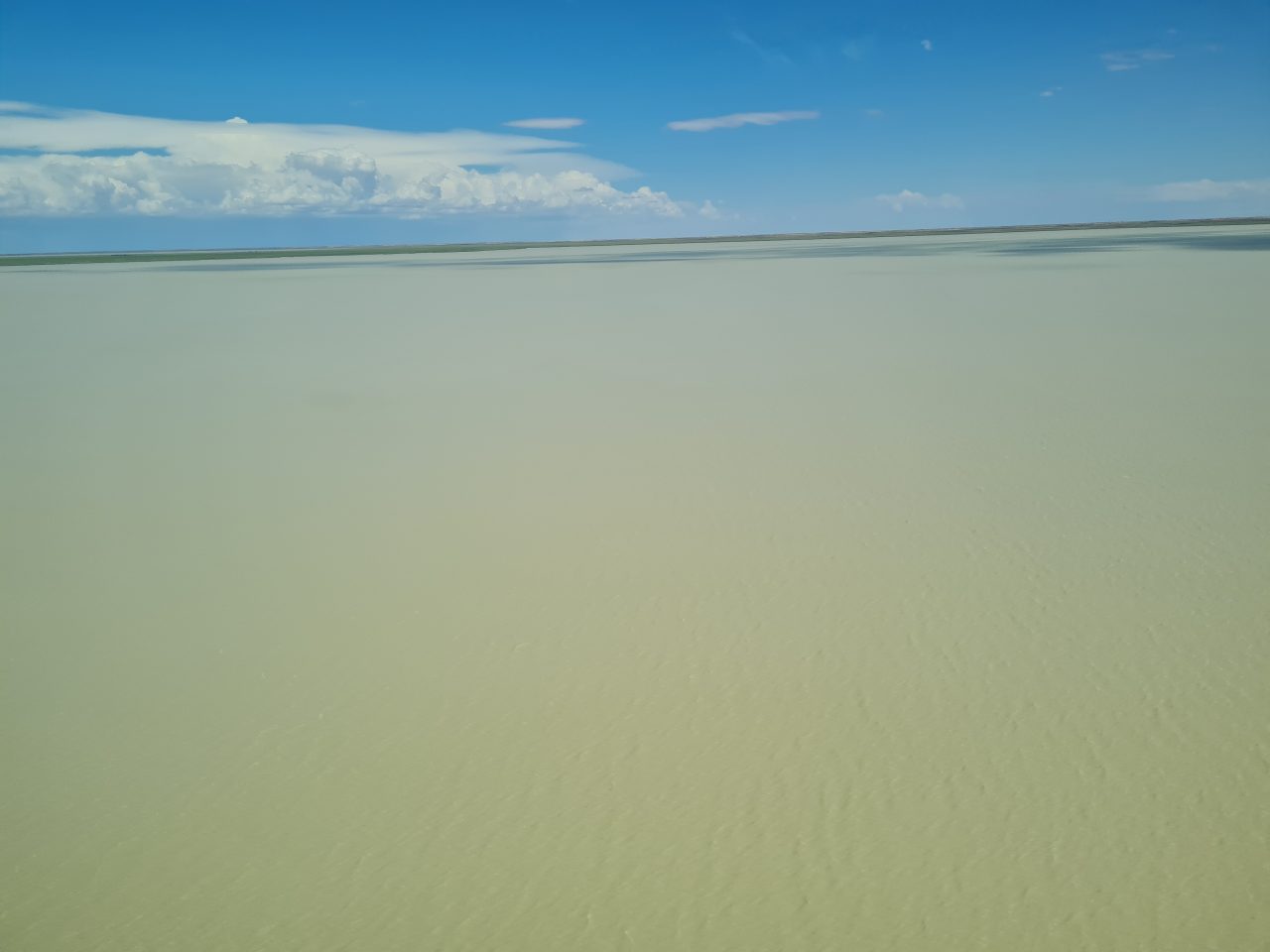 Aerial photo of large lake, beige water surface covering most of image  with a small band of land at the horizon. Sky is blue and clouds are high, white but building up.