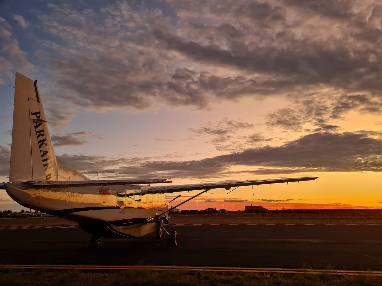 Photo of small cessna light aircraft on the tarmac, with a golden sunset  lighting up the horizon and reflecting in a warm yellow band along the length of the plane.