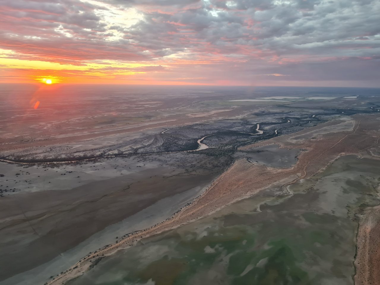 Aerial photo of river meandering across the floodplain at sunrise. Sky is orange and pink with cloud cover. 
