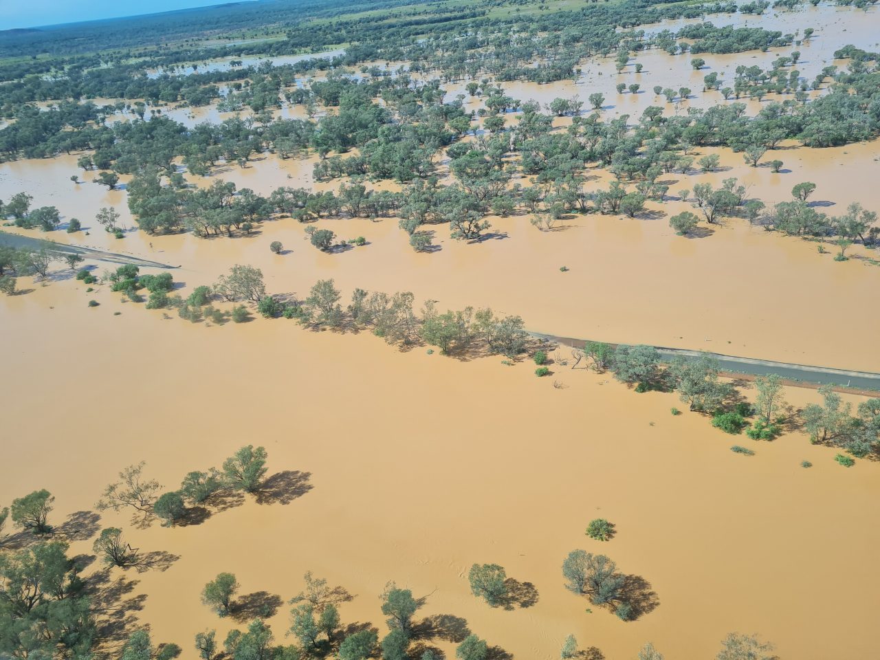 Aerial photo of floodwaters inundating the landscape with orange-brown water and submerging a section of sealed road making passing impossible . Trees on the floodplain are also in the expansive flood waters