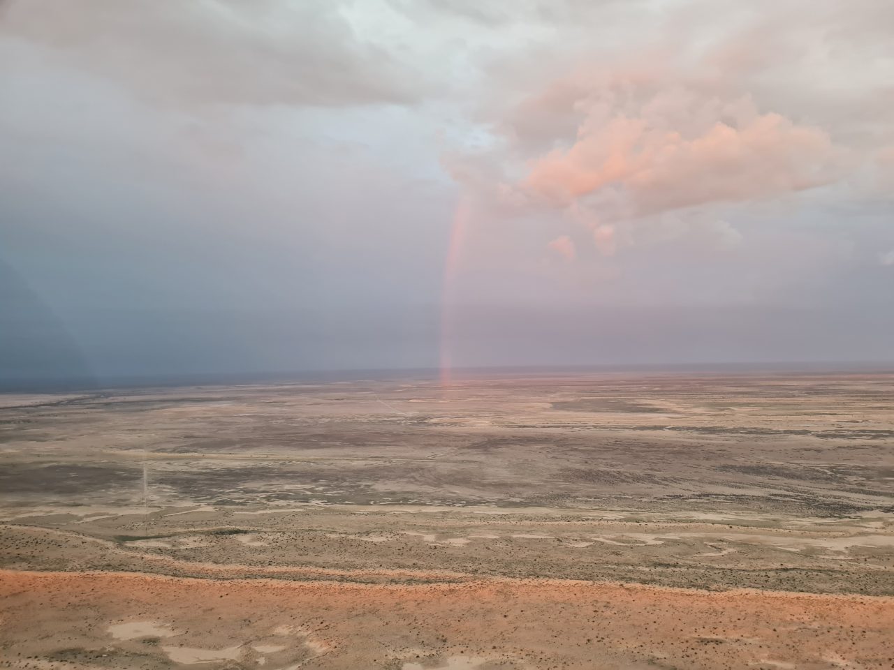 Aerial photo of landscape and sky. Landscape is expansive floodplain, sparsely vegetated and light yellow to orange coloured soils. Clouds are low, orange tinted and start of rainbow is visible.