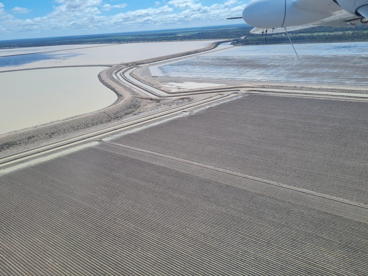 Aerial photo of landscape covered by large man made water storages that are rectangular in shape, divided by man made banks and full of beige coloured water. Over the banks are fields ploughed into giant corduroy texture and  waiting for planting.