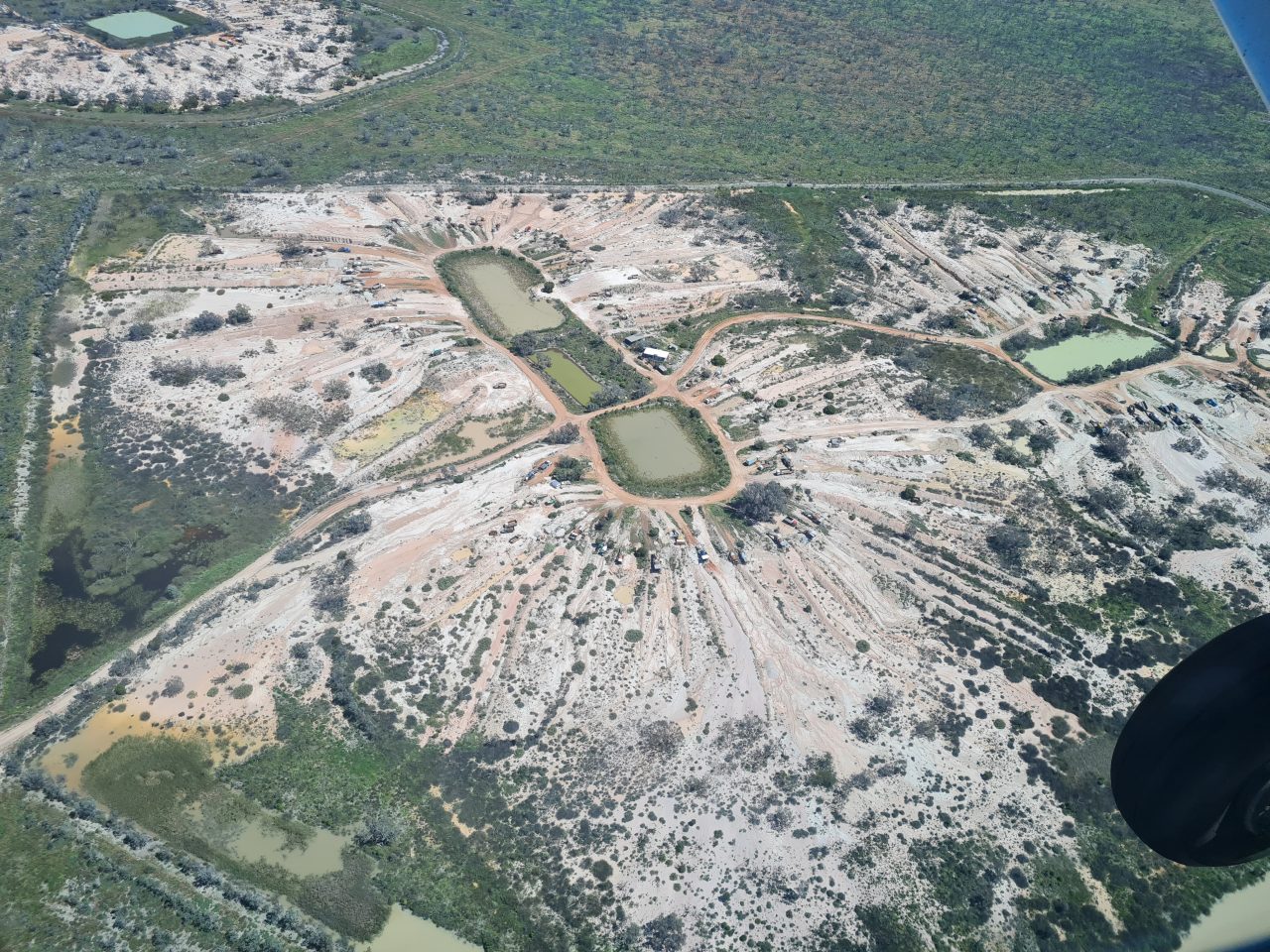 Aerial photo of cleared land surrounding rectangular dams with green water. Trucks parked in a ring around the dams with many dirt roads running from the dams out to the edge of the cleared areas. 