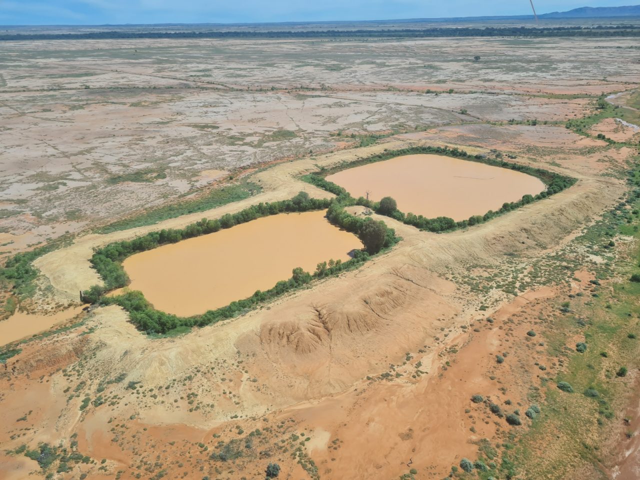 Aerial photo of two small rectangle farm dams holding orange-brown water in a desert landscape . Trees  and vegetation are growing in  thin line along the edge op the dams.  