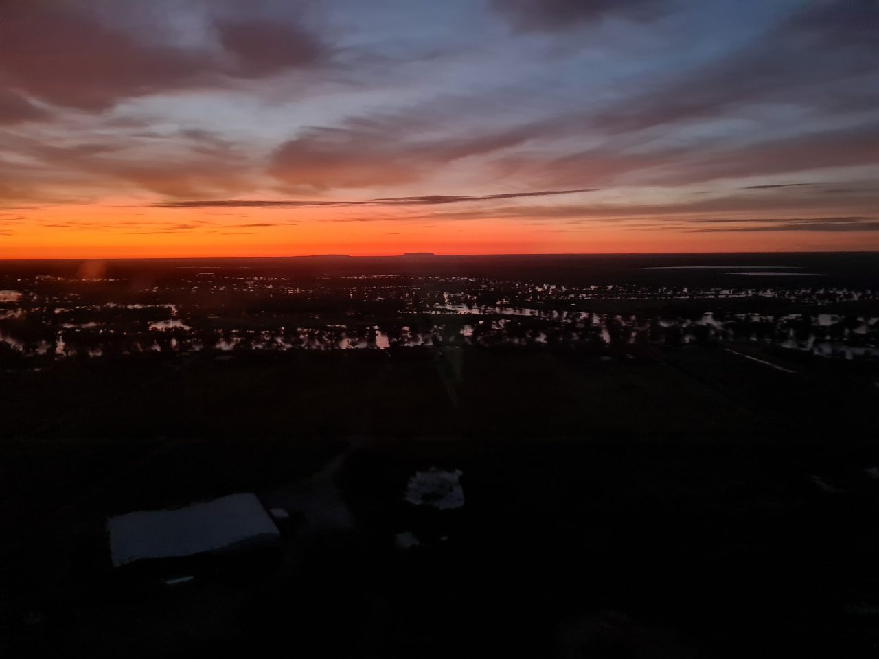 Aerial photo of sunrise over a river that is flooding with water inundating trees. The water is light coloured from the reflection of the sky. The horizon is specatualr with red and orange colours the foreground is dark in contrast