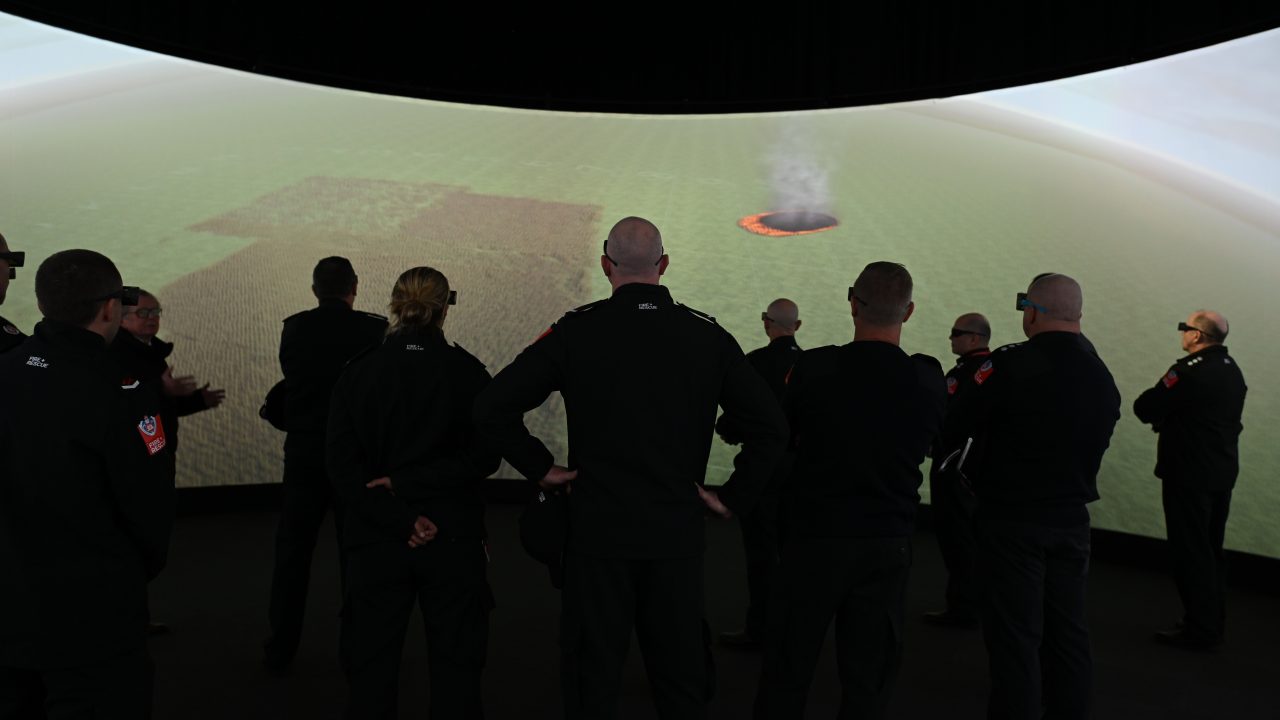 Photograph of a group of firefighters facing a cinematic screen displaying a green landscape with a fire starting on screen