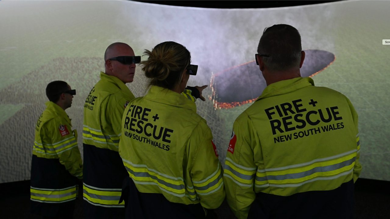 Image of four firefighters viewing a fire simulation on an immersive cinema screen facing away from the camera