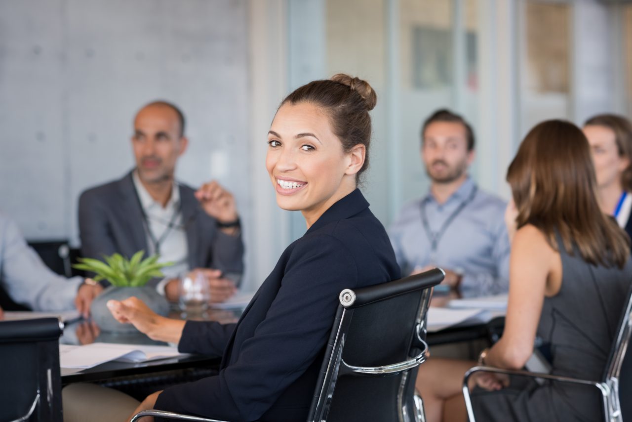 Young beautiful businesswoman with colleagues sitting in a modern board room. Proud smiling business woman sitting during a meeting and looking at camera. Portrait of a happy businesswoman with executives working.