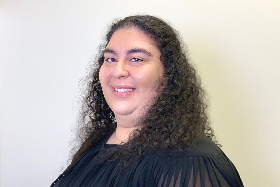 Dr. Anthea Charalambous with long curly hair, smiling warmly and wearing a black pleated top, standing in front of a neutral wall.