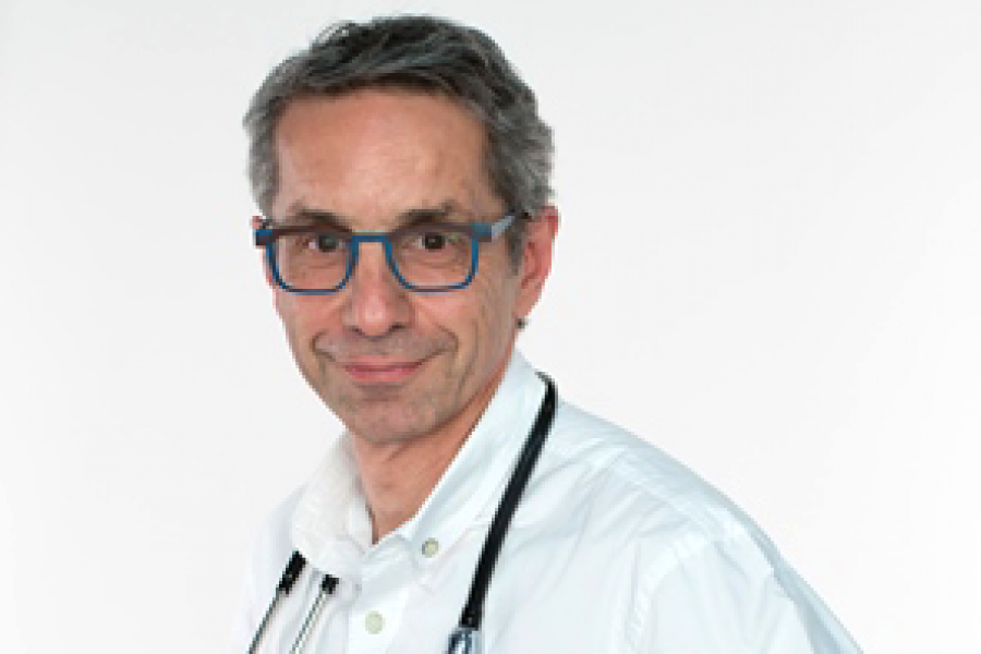 Older man with short gray hair and blue-framed glasses, dressed in a white coat with a stethoscope draped around his neck, posing against a white background.