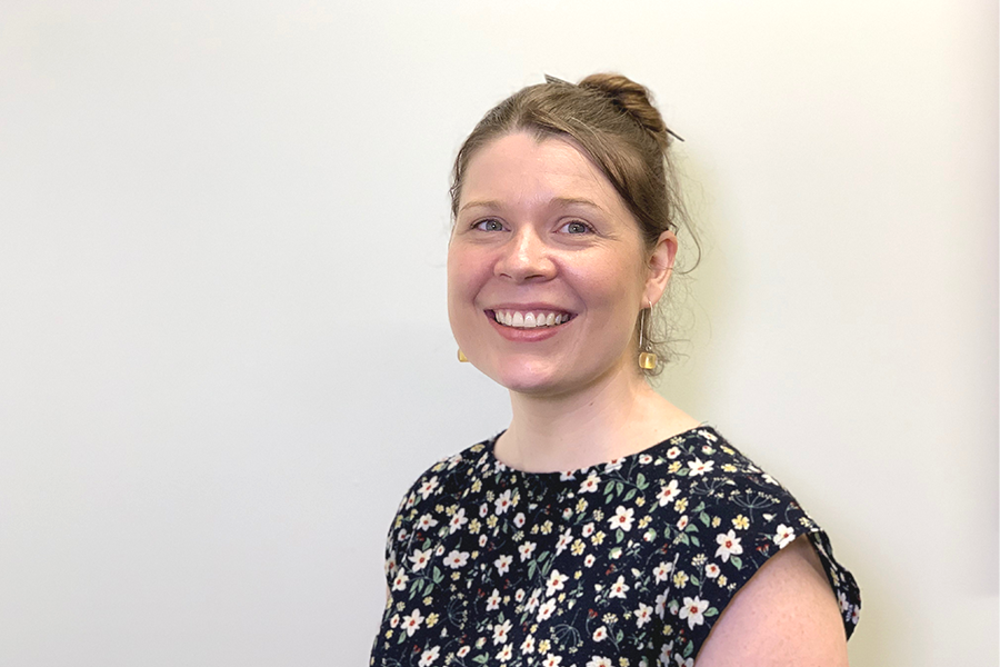 Smiling Dr. Crystal McKeough in a black floral blouse with hair tied back, standing against a pale background and wearing small dangling earrings.