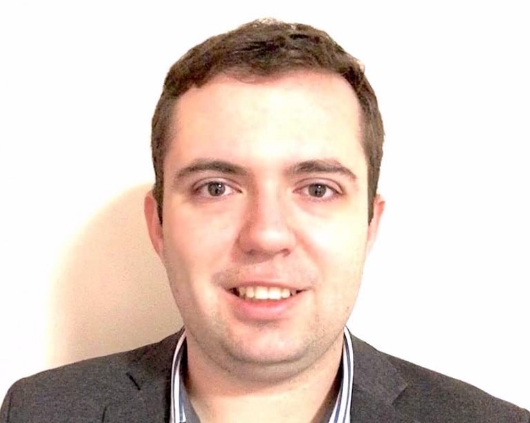 Portrait of a Dr Michael Petinga in a grey blazer over a dark sweater and collared shirt, standing in front of a plain white background with a friendly expression.