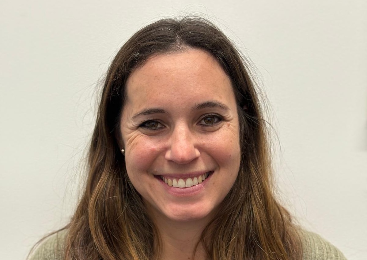 Informal portrait of Dr. Monique Akouri with long brown hair, wearing a green knit cardigan and a silver necklace, in front of a white wall.