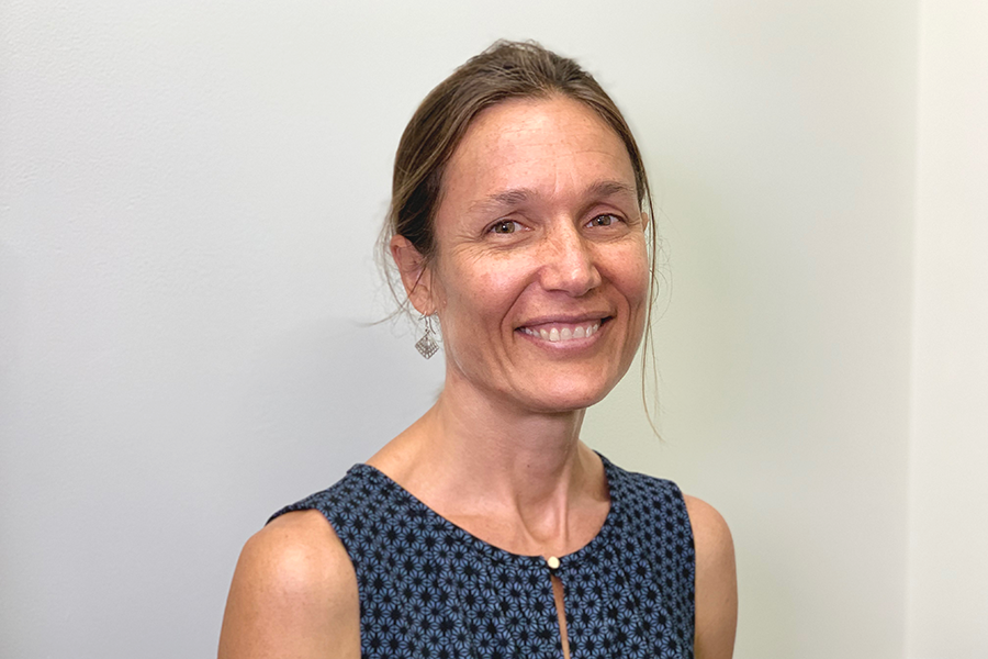 Dr. Sarah Brewster with tied-back hair wearing a blue patterned sleeveless top, smiling gently in a softly lit room.