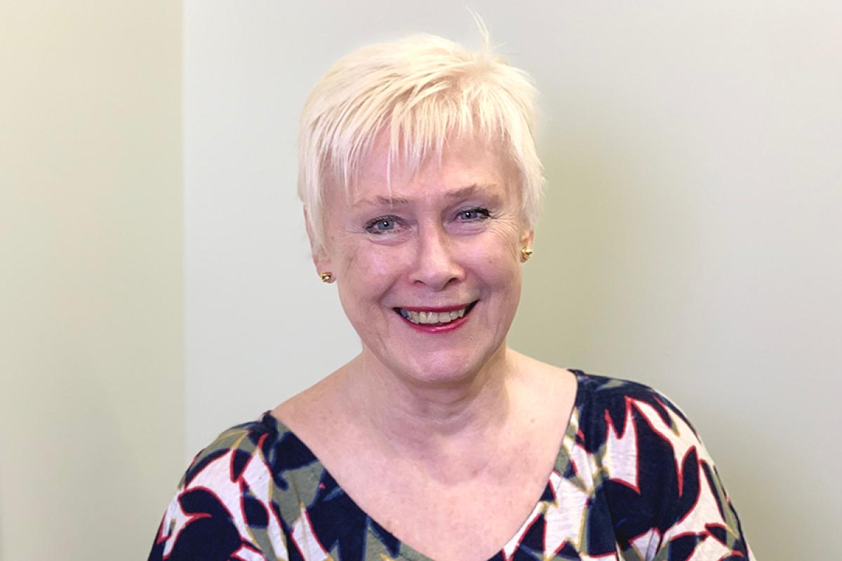 Portrait of Dr. Mary Jurek with short platinum blonde hair, wearing a patterned dress with red, white, and green accents, posed against a pale neutral background.